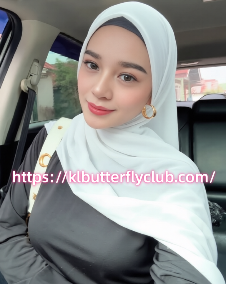 Portrait of a woman in a light blue hijab with gold hoops, sitting in a car and smiling at the camera; pink URL watermark across the image.
