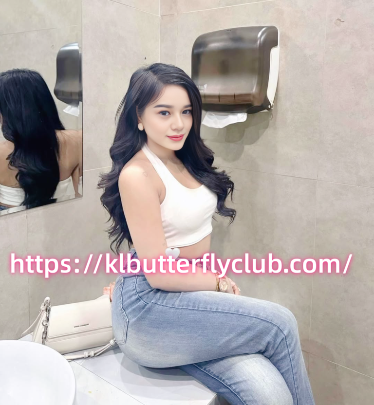 Young woman in a white sleeveless top and light jeans sits on a bathroom counter, looking at the camera, with a pink URL across the image.