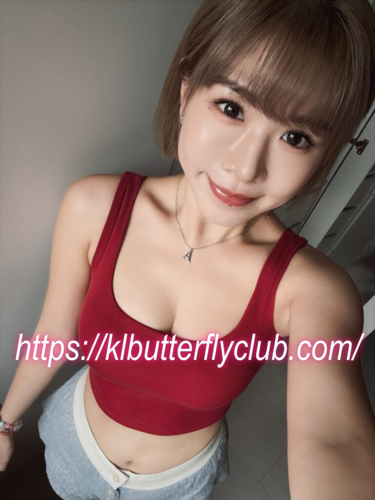 Close-up selfie of a young woman with short blond hair wearing a red sleeveless top, necklace with an A pendant, and light blue jeans; pink lipstick; a URL overlay reads 'https://klbutterflyclub.com/'
